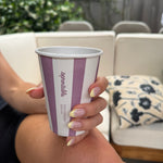 A person holding a compostable coffee cup with purple and white vertical stripes and the brand name 'Sproutable' printed on it, set against an outdoor patio background with a white sectional sofa.