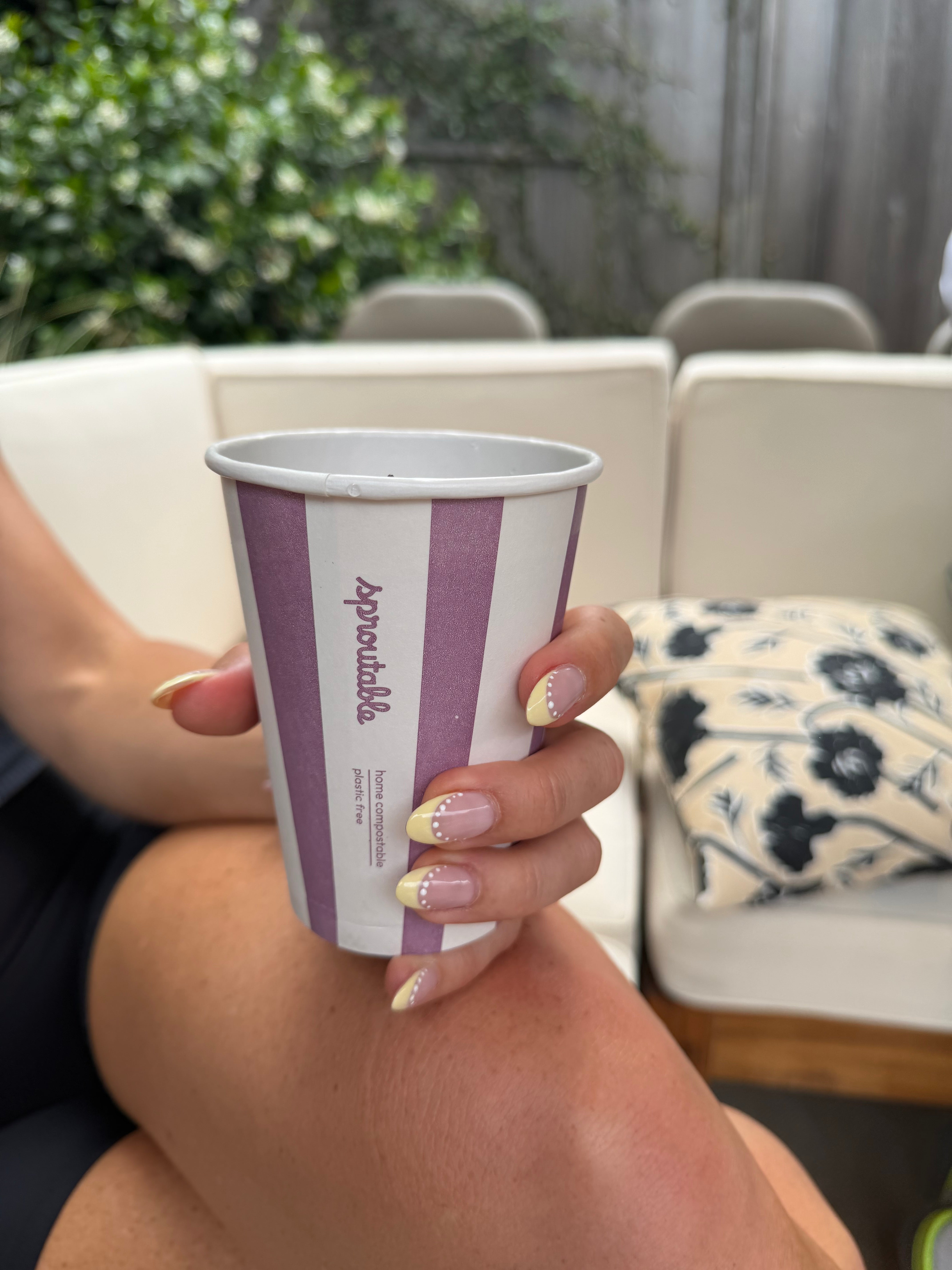 A person holding a compostable coffee cup with purple and white vertical stripes and the brand name 'Sproutable' printed on it, set against an outdoor patio background with a white sectional sofa.